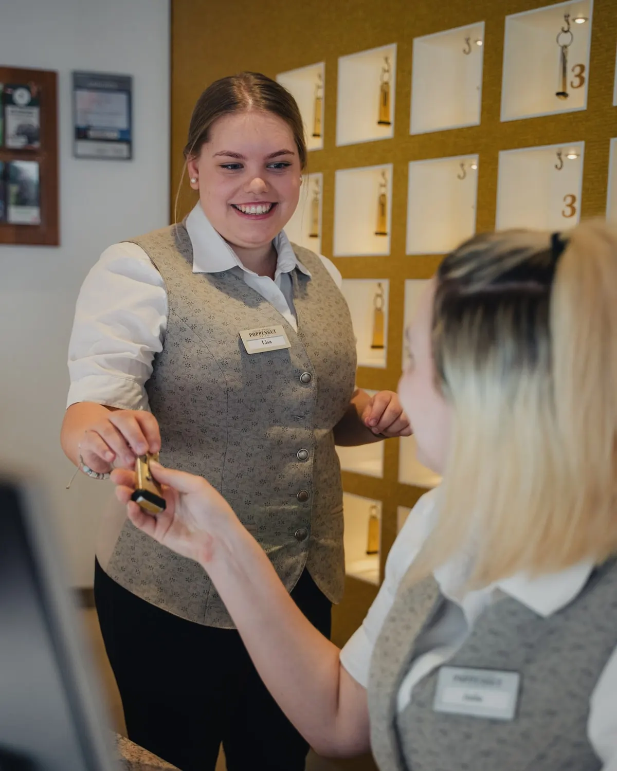 Pracovnice recepce se smíchem podává klíč od pokoje, za ní tabule s klíči