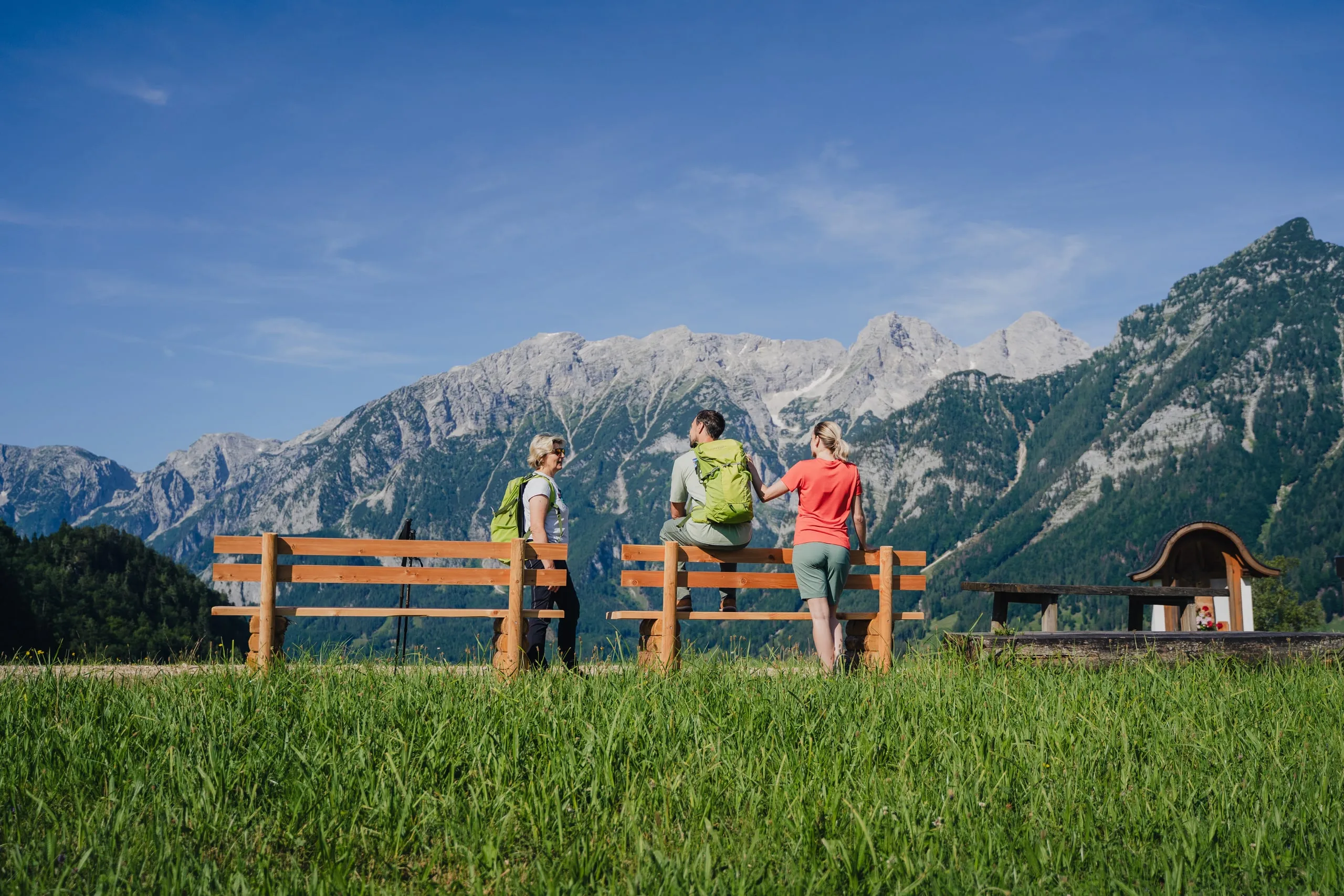 Tři turisté u dřevěné lavice na horské louce, za nimi hory