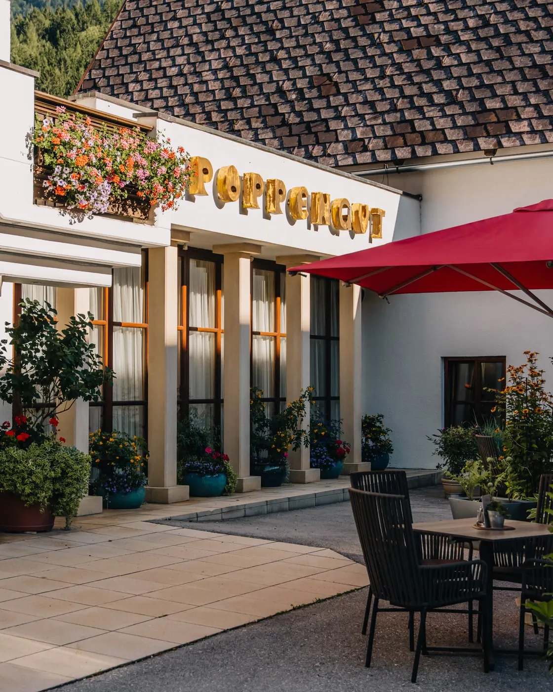 © Hartwig Winkler Vchod do hotelu Poppengut v létě, zlatý nápis nad vstupem, truhlík s květinami, červený slunečník a stůl na terase