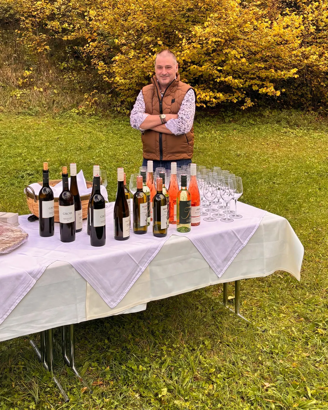 © Hartwig Winkler Muž v hnědé vestě stojí za bíle prostřeným stolem s lahvemi vína a skleničkami na louce, za ním žluté podzimní listí