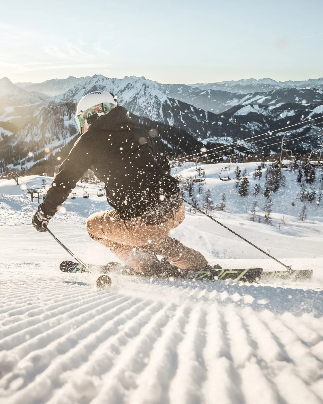 © Moritz Ablinger / Oberösterreich Tourismus GmbH Lyžař krájí oblouk na čerstvě upravené sjezdovce proti nízkému slunci, za ním zasněžené hory