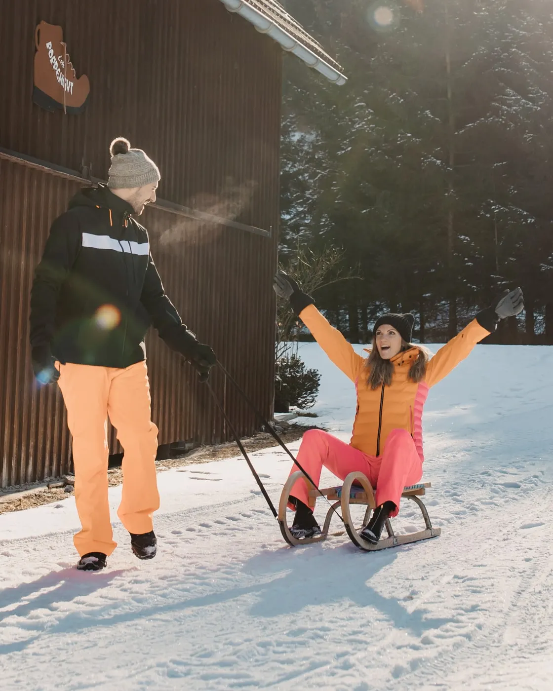 © Selina Flasch Photography Pár na sáňkách před dřevěnou stodolou ve sněhu: on táhne sáňky, ona na nich sedí a jásá se zdviženýma rukama