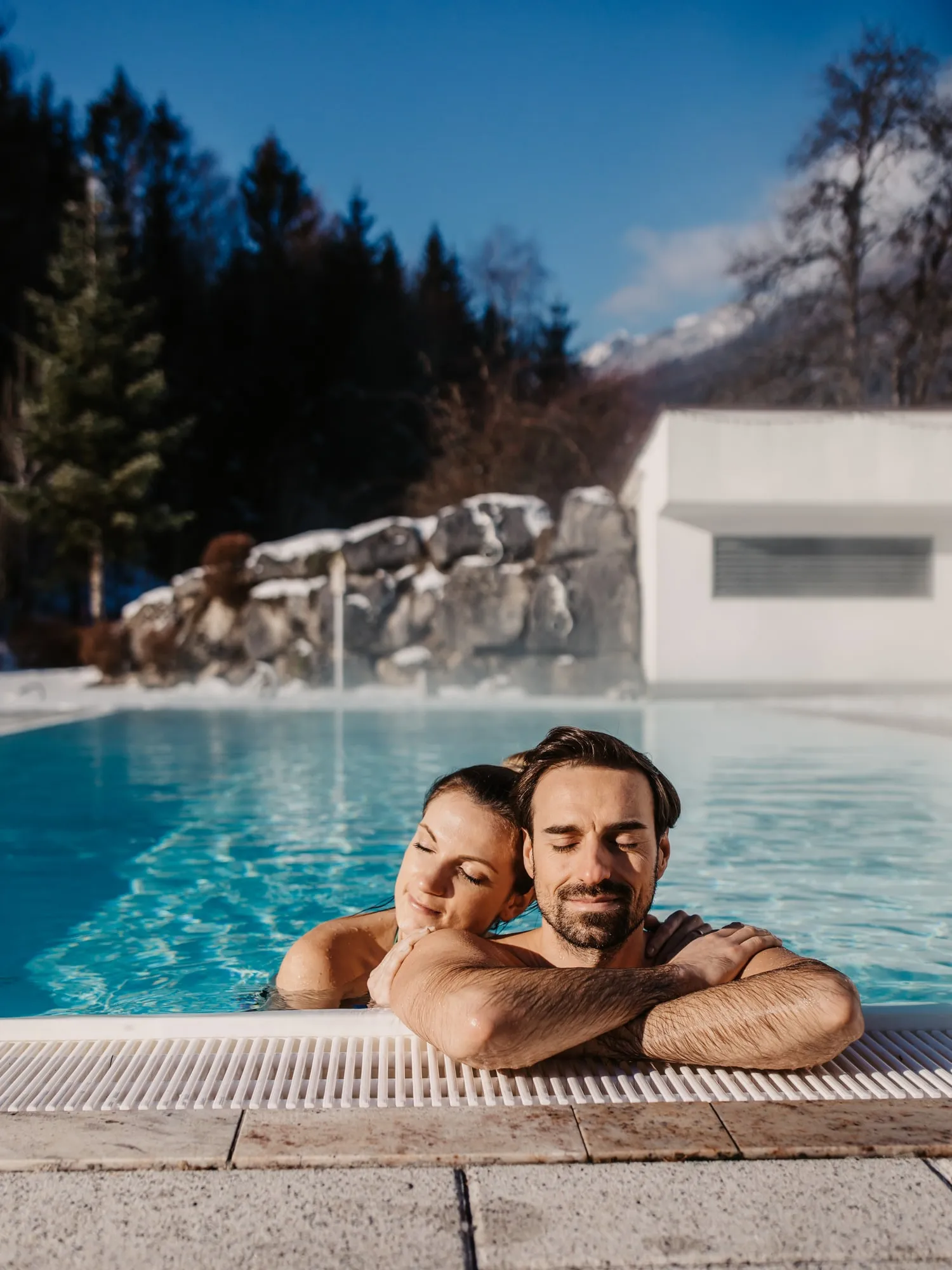 © Selina Flasch Photography Pár odpočívá na okraji teplého venkovního bazénu, za nimi zasněžené stromy