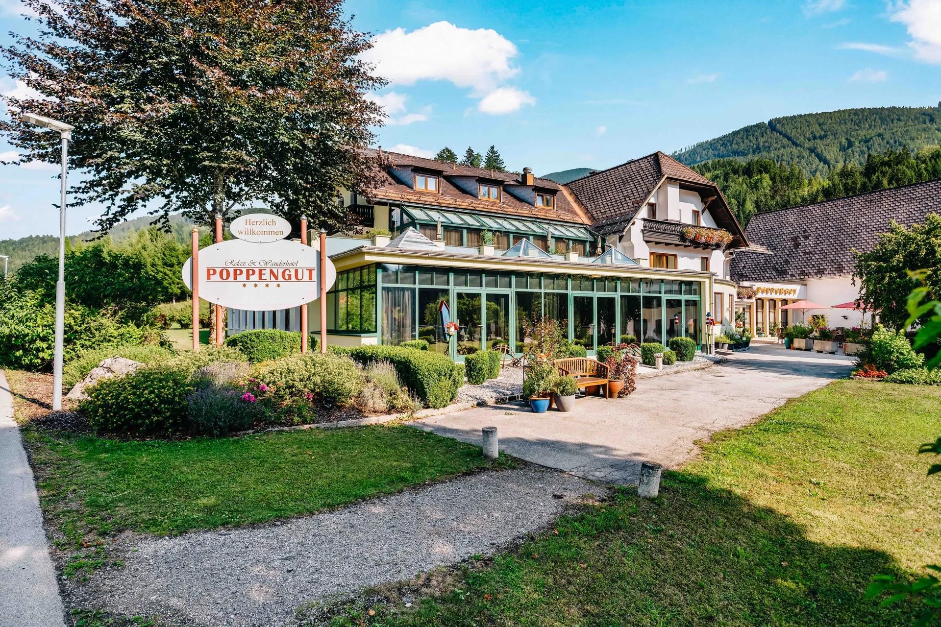 © Hartwig Winkler Vchod do hotelu Poppengut v létě – tabule „Herzlich willkommen“, zimní zahrada a za ní dům