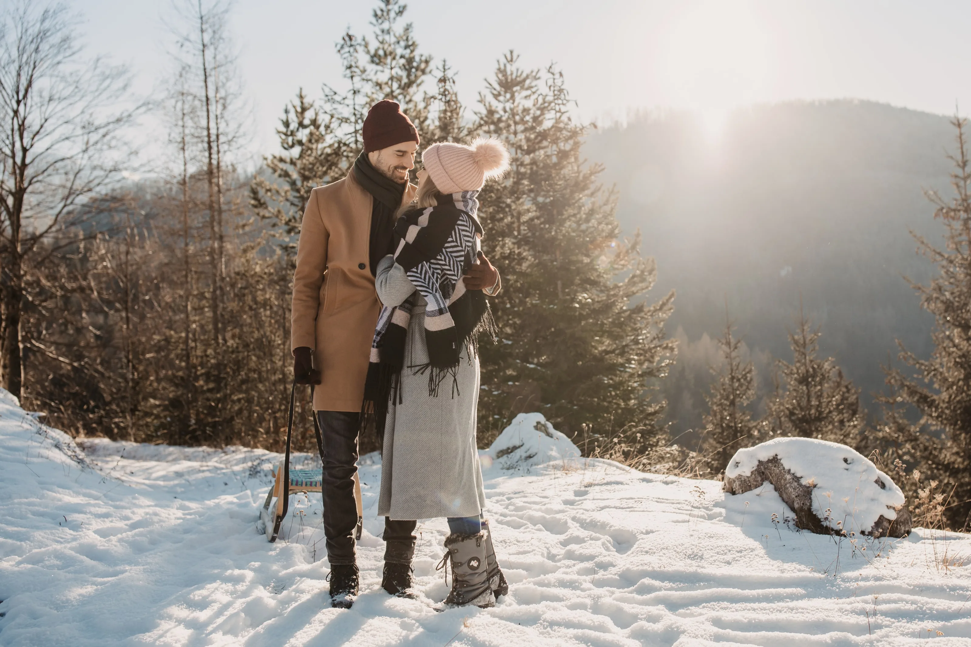 © Selina Flasch Photography Pár stojí s dřevěnými sáněmi ve sněhu, za nimi lesnatý hřeben na slunci