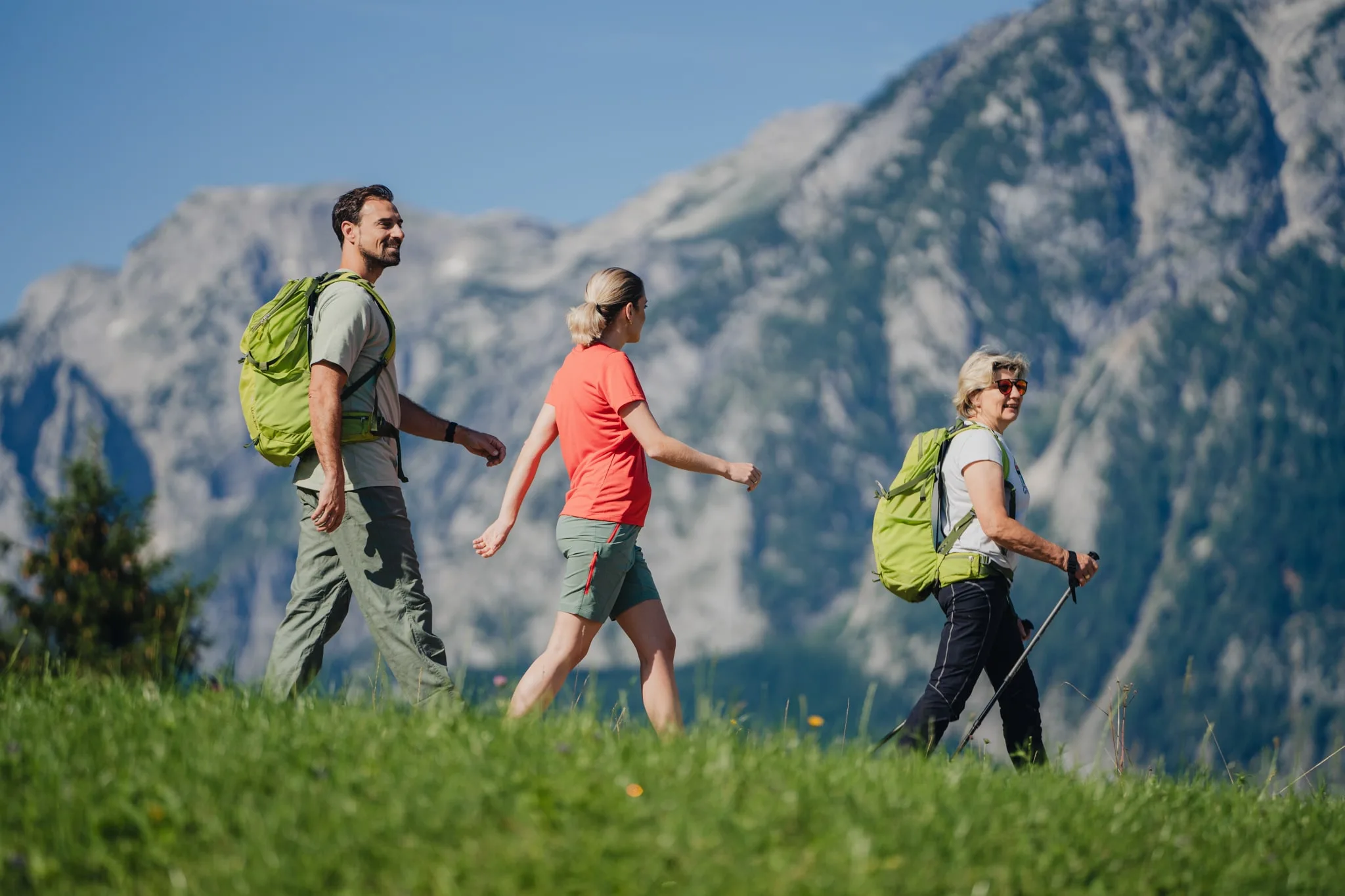 Tři turisté s batohy jdou po horské louce, za nimi se zvedá skalnatý vrchol