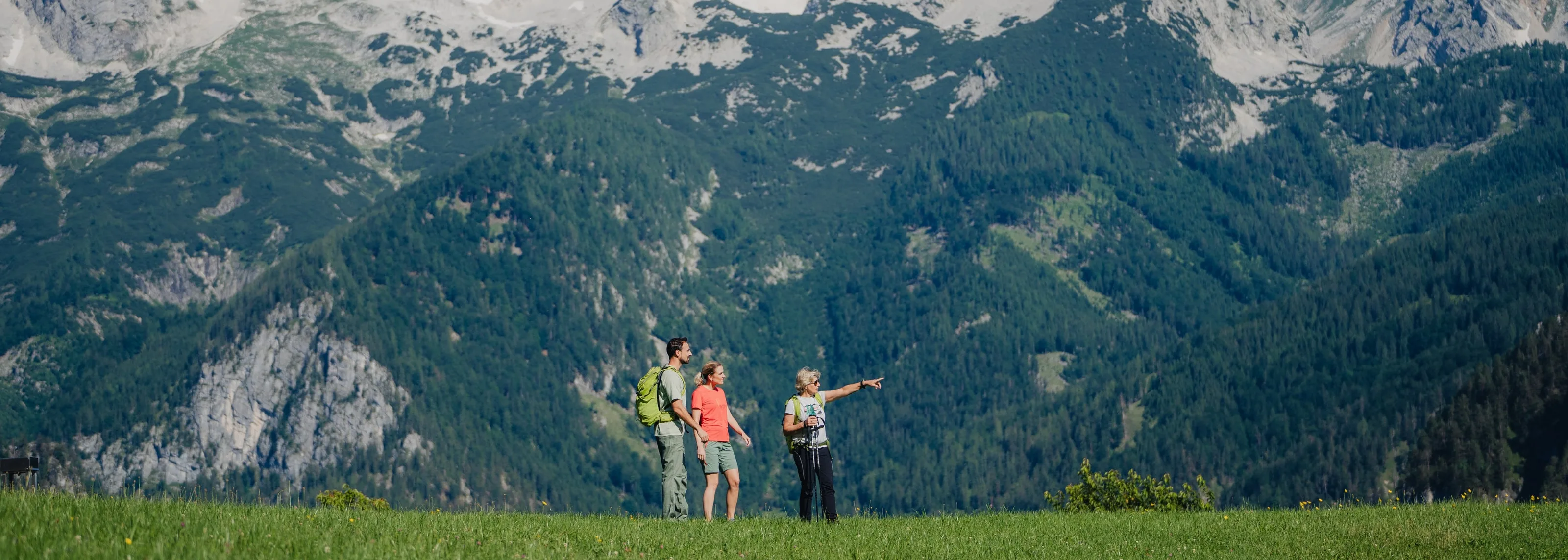 © Sarah Kreutzhuber Drei Wanderer auf einer Wiese, eine zeigt den Weg – dahinter die bewaldeten Hänge und Felsen des Toten Gebirges